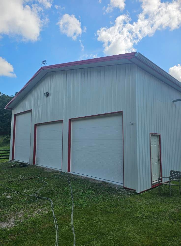 White metal garage building with three roll-up doors and a side entry door on a grassy lot under a cloudy sky