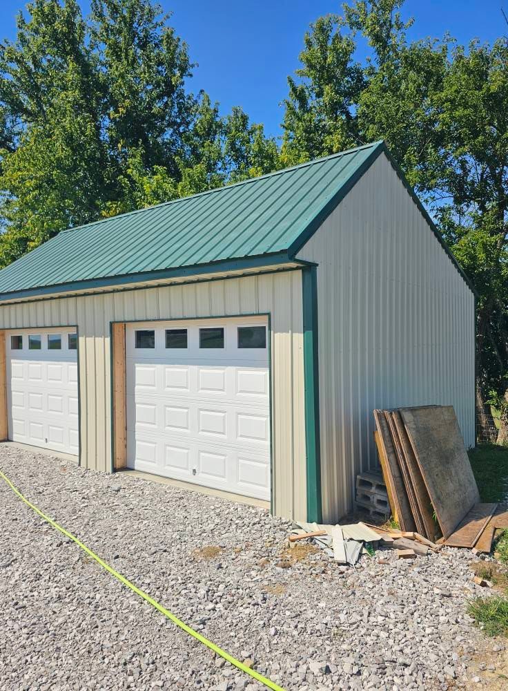 Two-car detached garage with green metal roof and white doors on a gravel lot, with boards leaning against it.