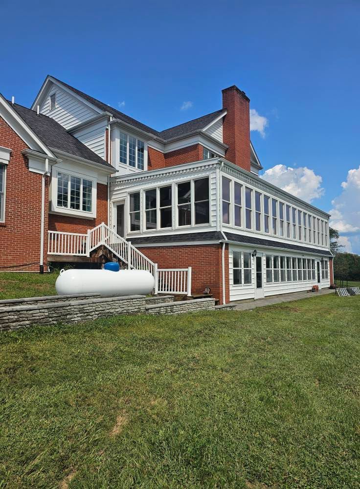 Brick house with white enclosed porch, red chimney, and propane tank on a grassy lawn under blue sky