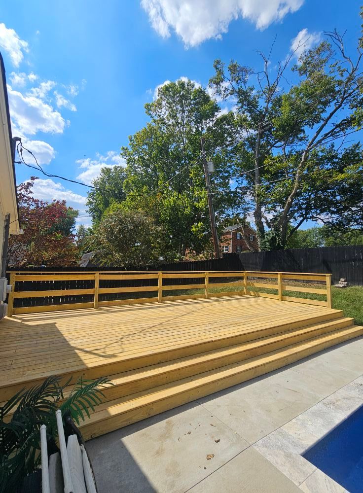 Wooden deck with steps beside a building, bordered by a railing and trees under a blue sky.