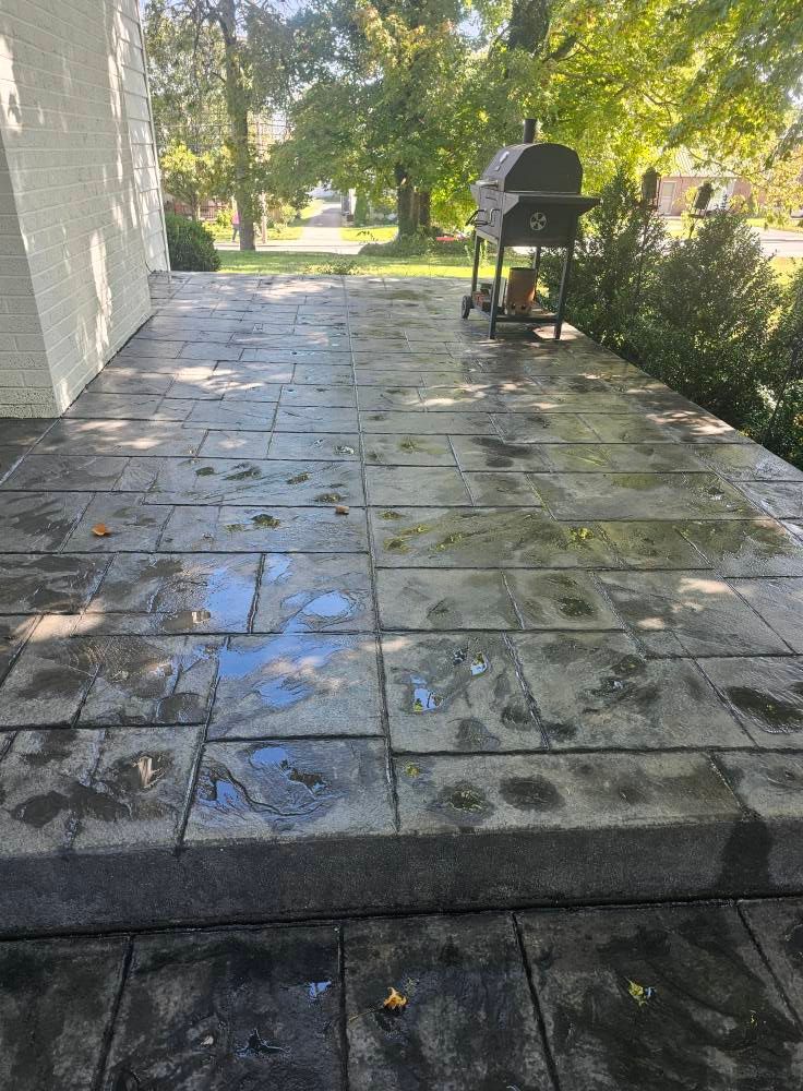 Stone patio walkway beside a house, with a barbecue grill and hedges in the background.
