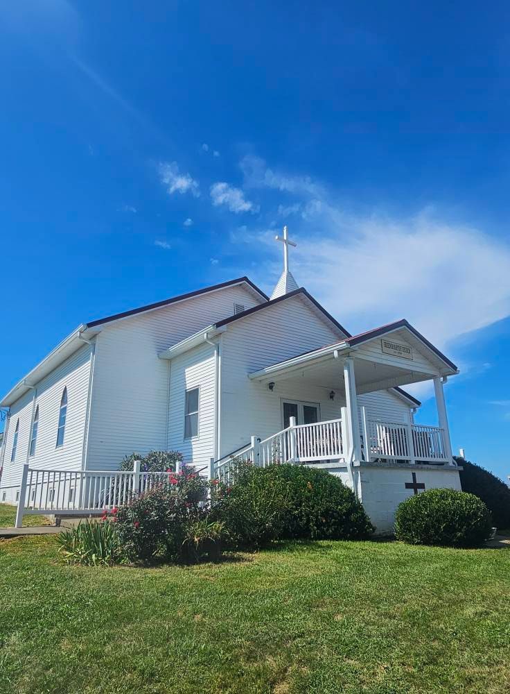 White church with steeple and porch on a grassy hill under a bright blue sky