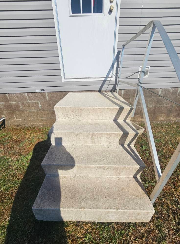 Concrete front steps leading to a white door, with a metal handrail and siding on the house.