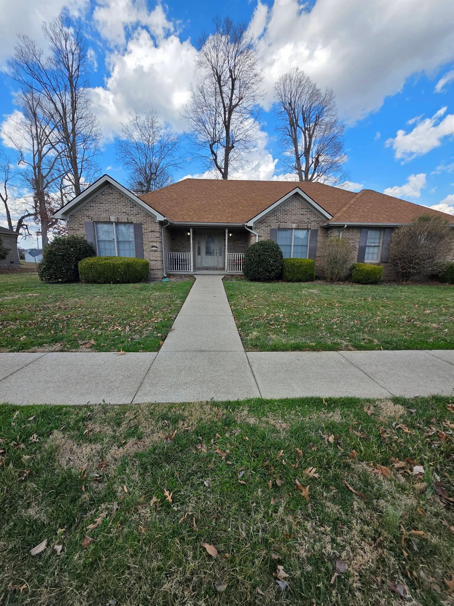 Single-story stone house with a red roof, front walkway, and leaf-covered lawn under a blue sky.