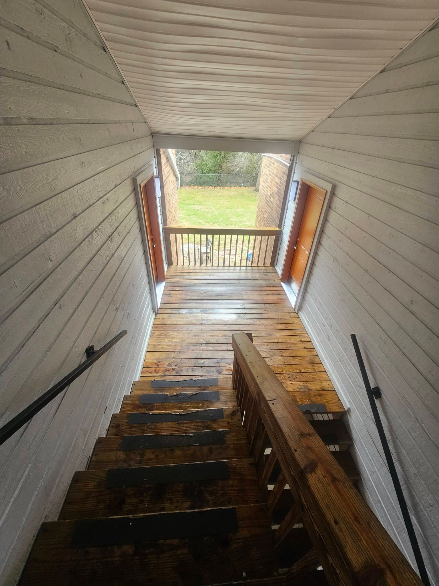 Narrow stairwell with worn wooden steps leading to a small porch and a view of greenery outside.