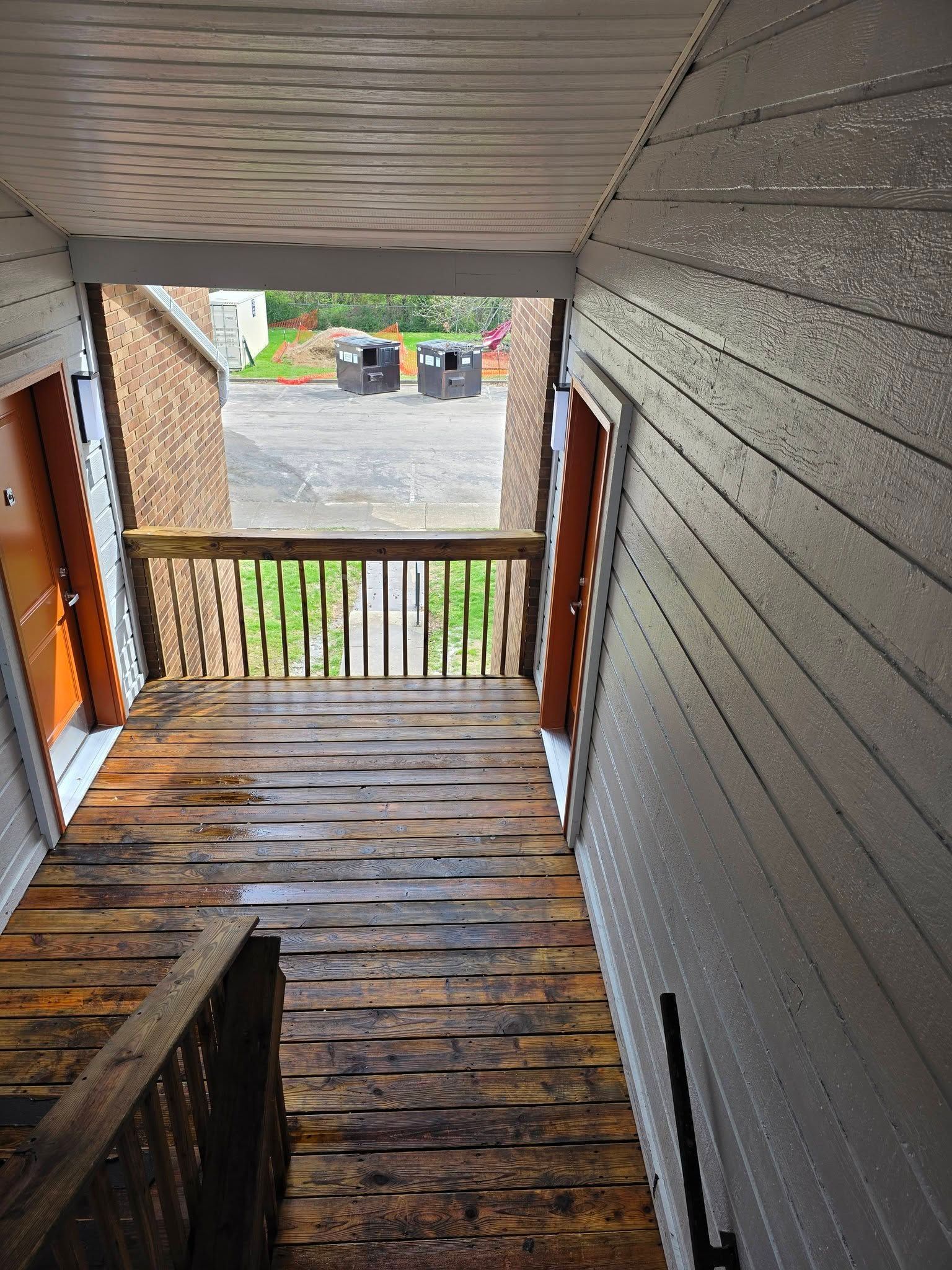 Narrow covered porch with worn wooden floorboards and railing, overlooking a gravel driveway and bins below