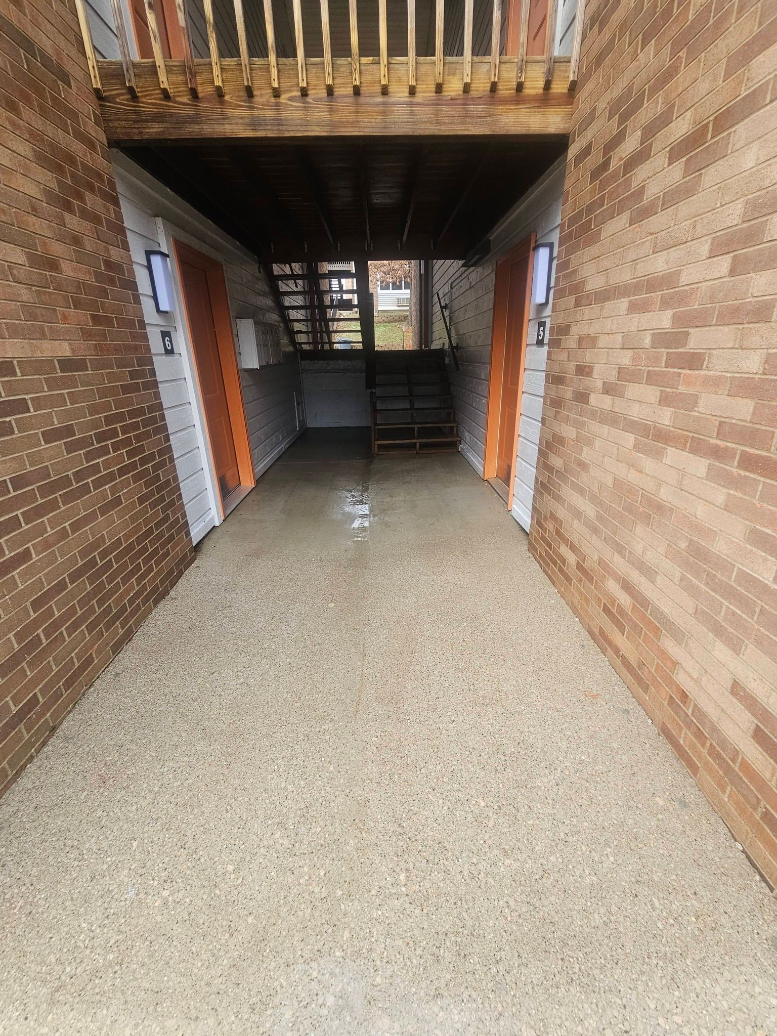 Brick-lined corridor with terrazzo floor and a balcony overhead, leading to a dark doorway at the end