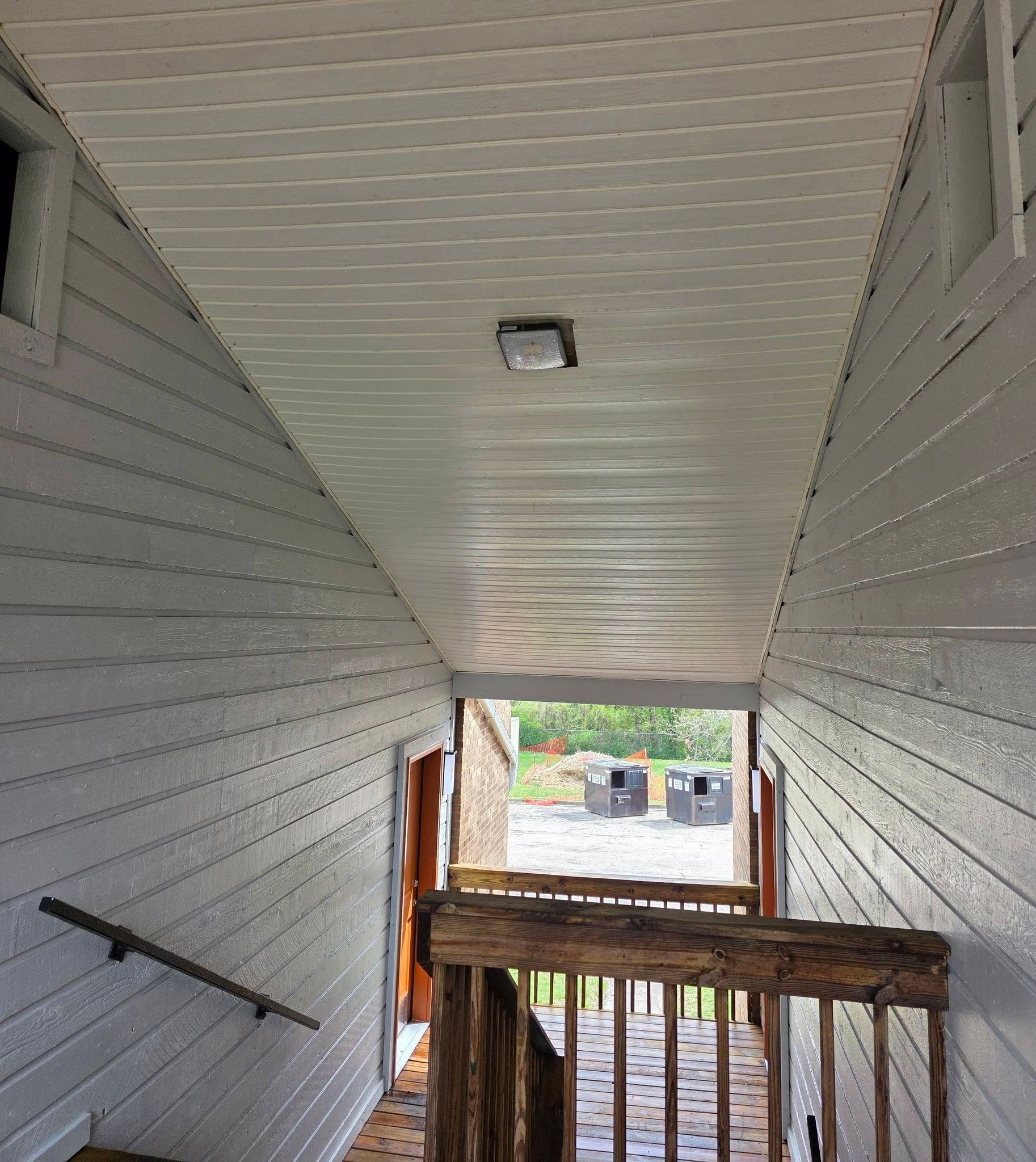 Covered outdoor stairwell with gray siding and a wooden railing, looking down to a yard below