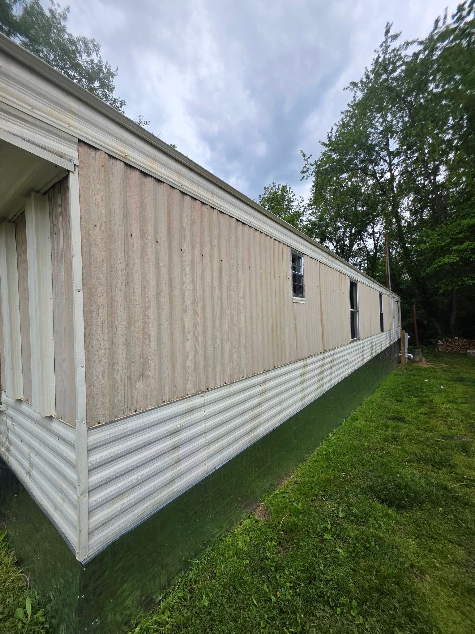 Long beige mobile home with white trim and skirting beside grass and trees under a cloudy sky