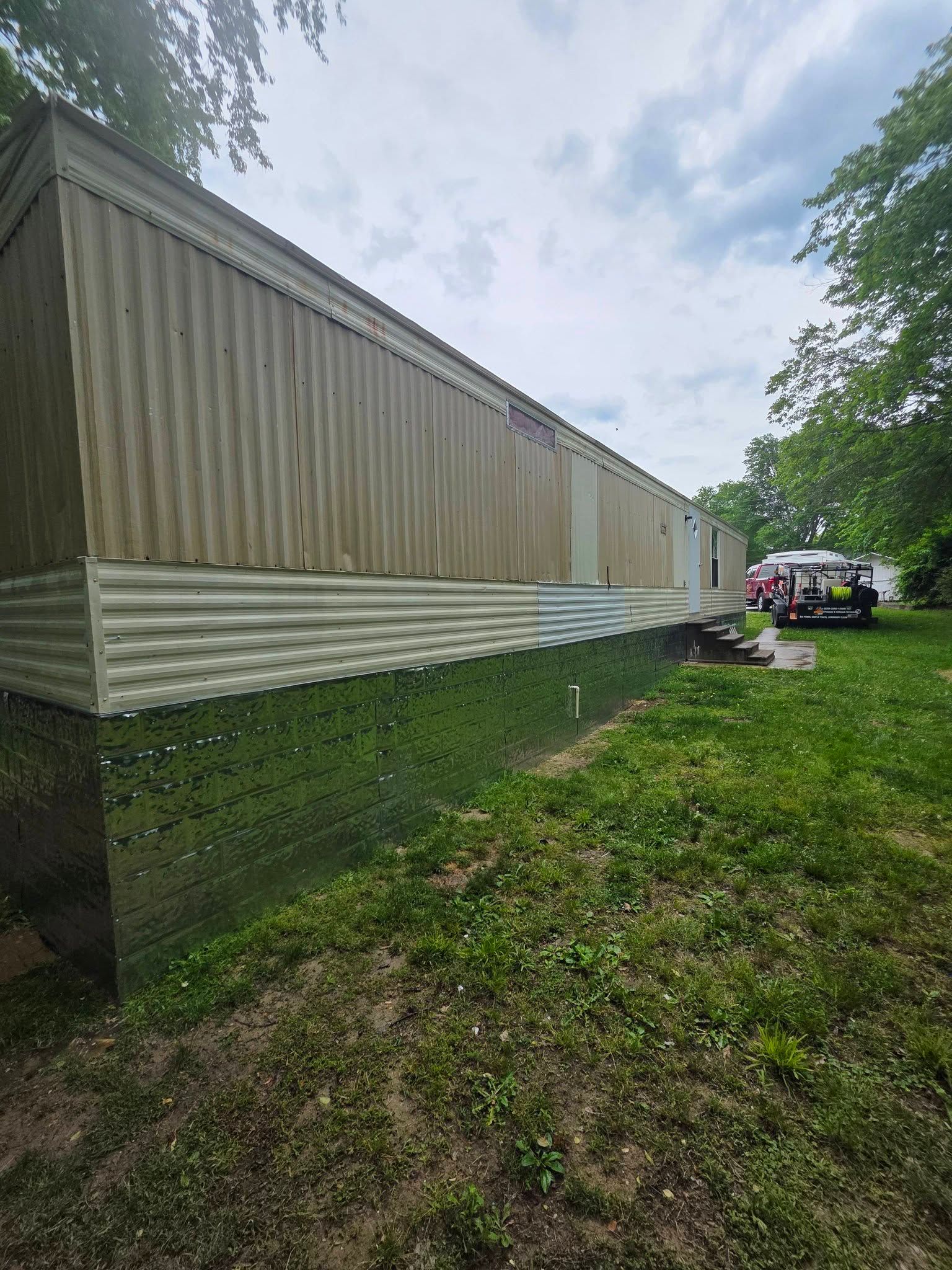 Long beige mobile home on a grassy lot beside trees and a utility vehicle in the distance.