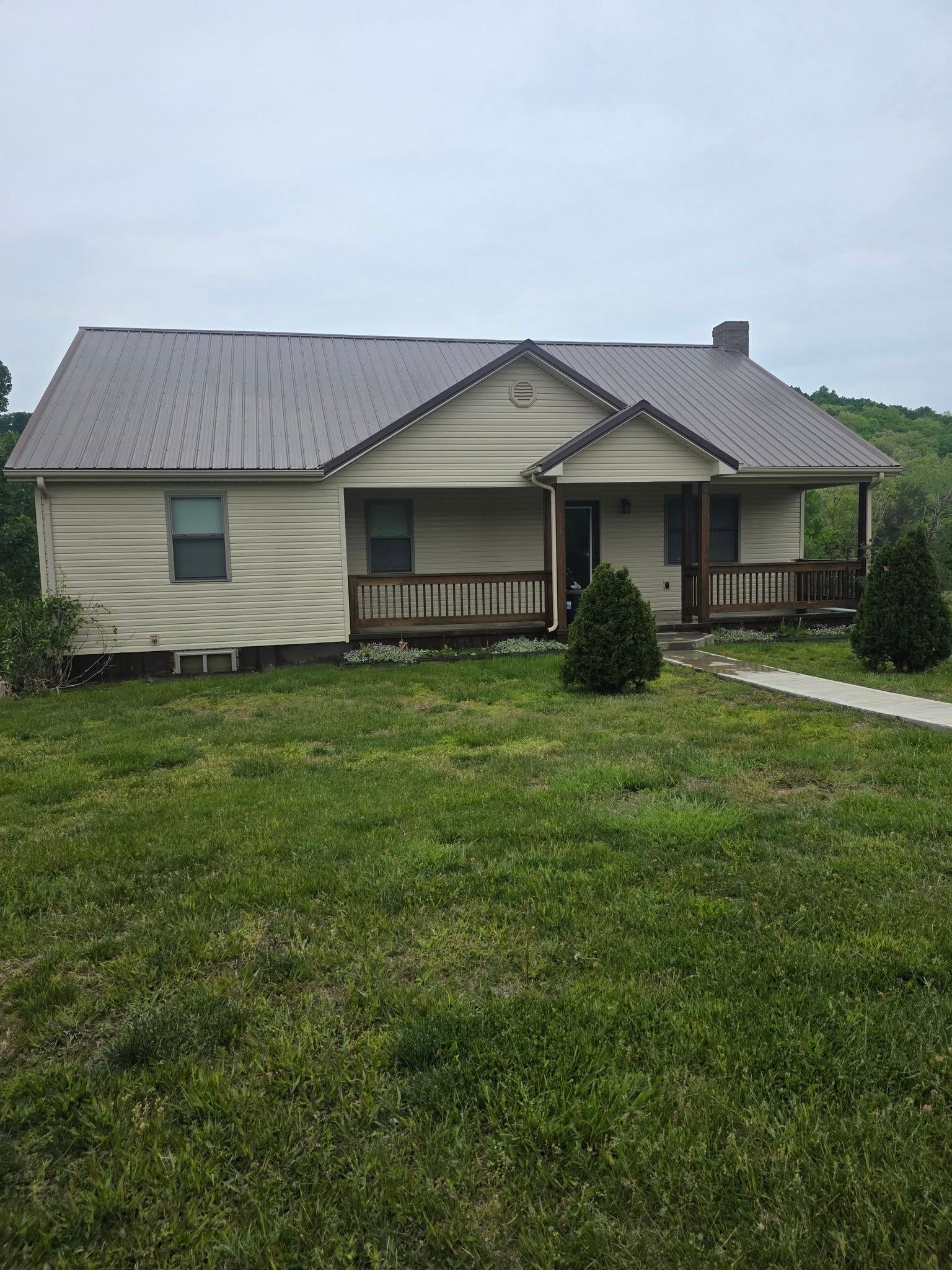 Small beige house with a gray roof, front porch, and a large grassy yard under an overcast sky