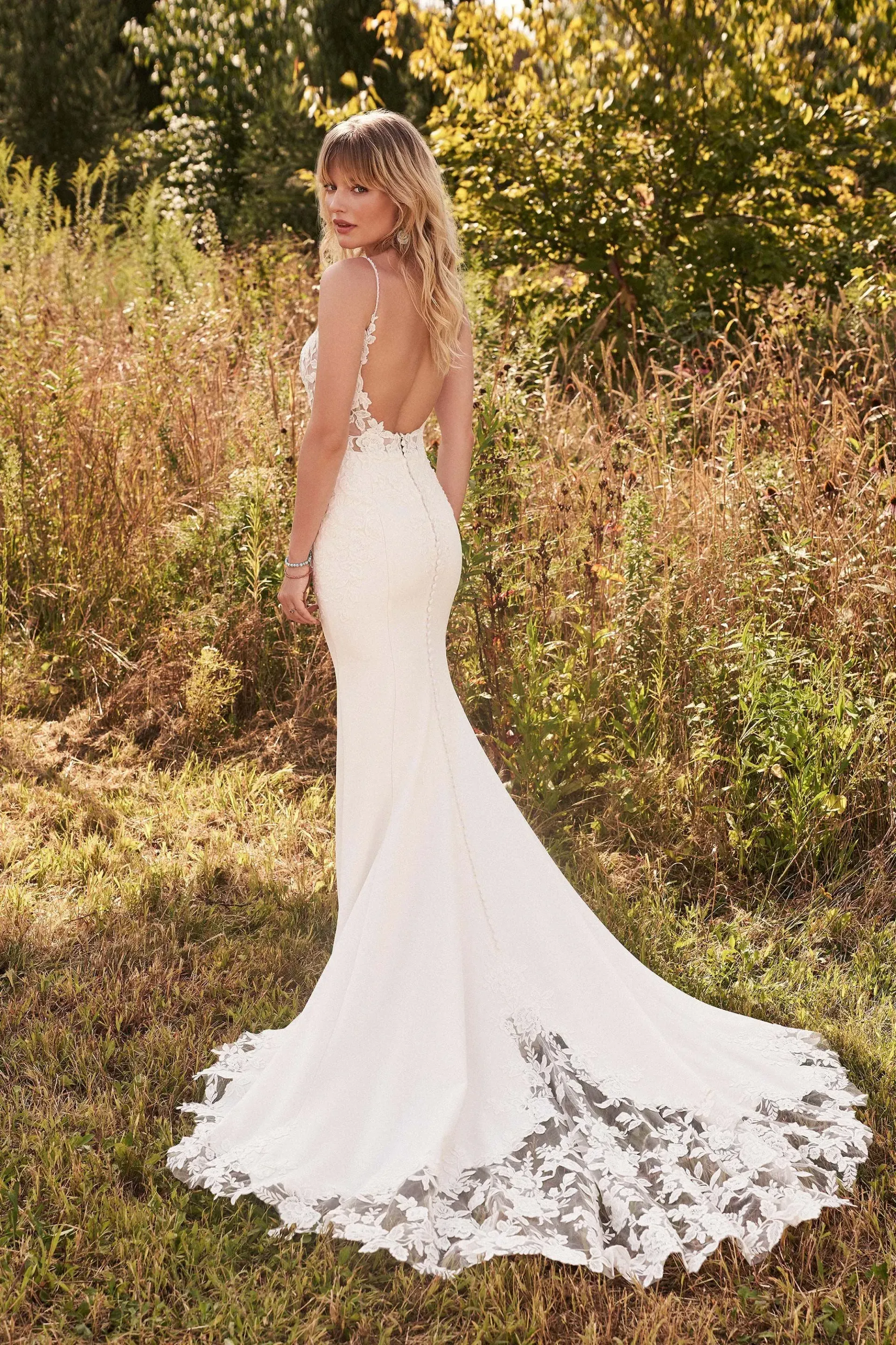 Woman in a white wedding dress with a long train, standing outdoors with a natural background.