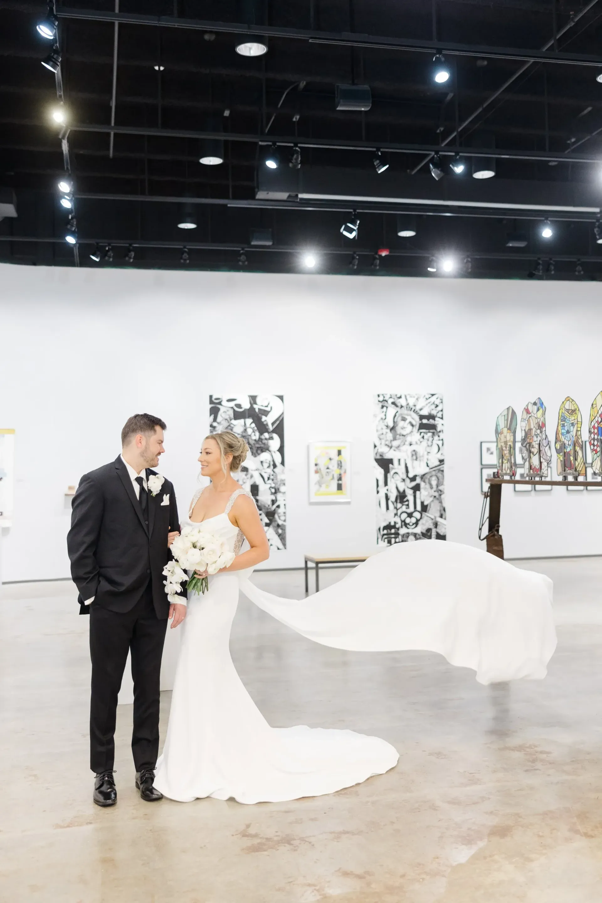Bride and groom pose in art gallery; bride's train flows behind her. They smile at each other.