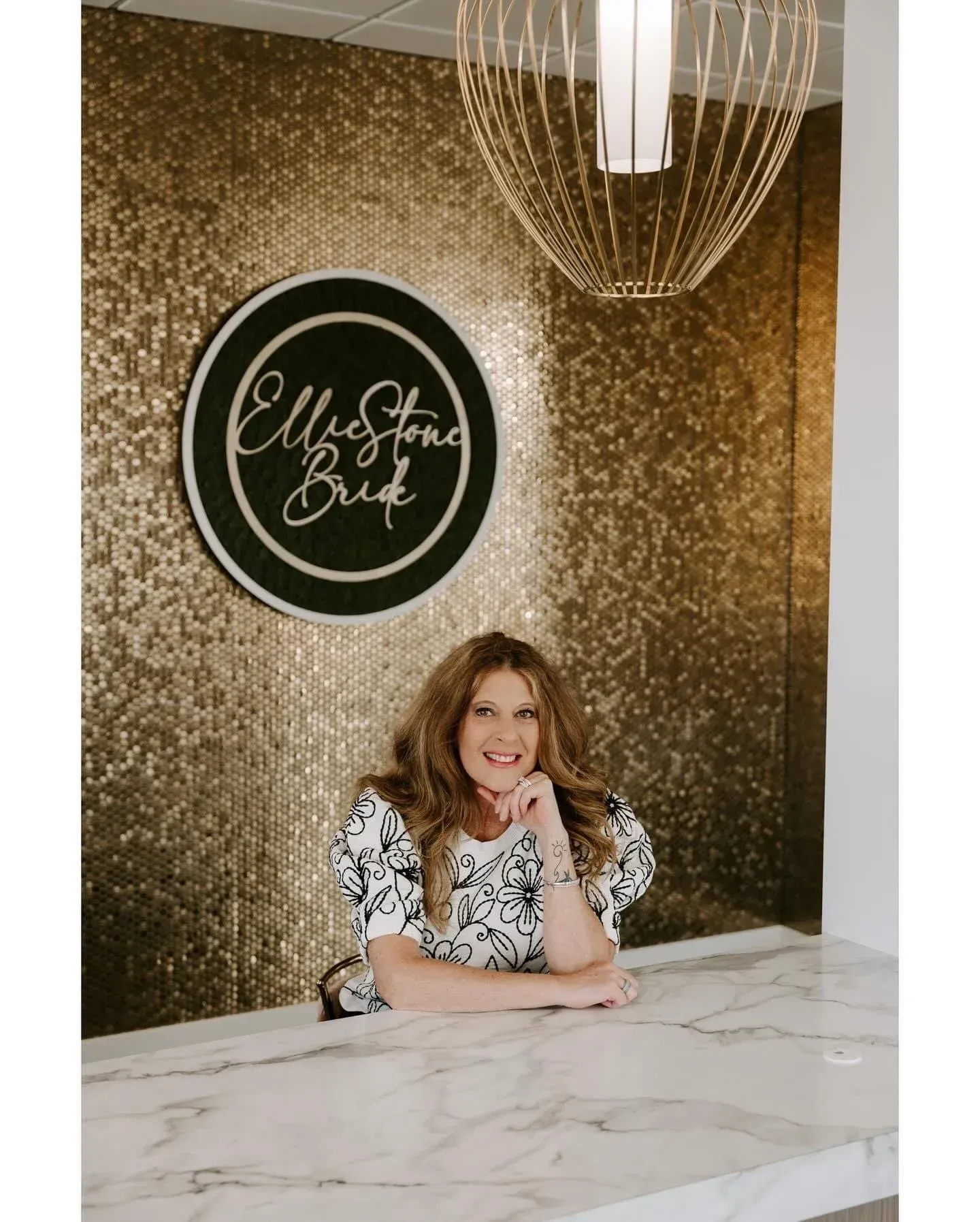 Woman at a white marble counter smiling, with a gold-tiled wall and a logo that says