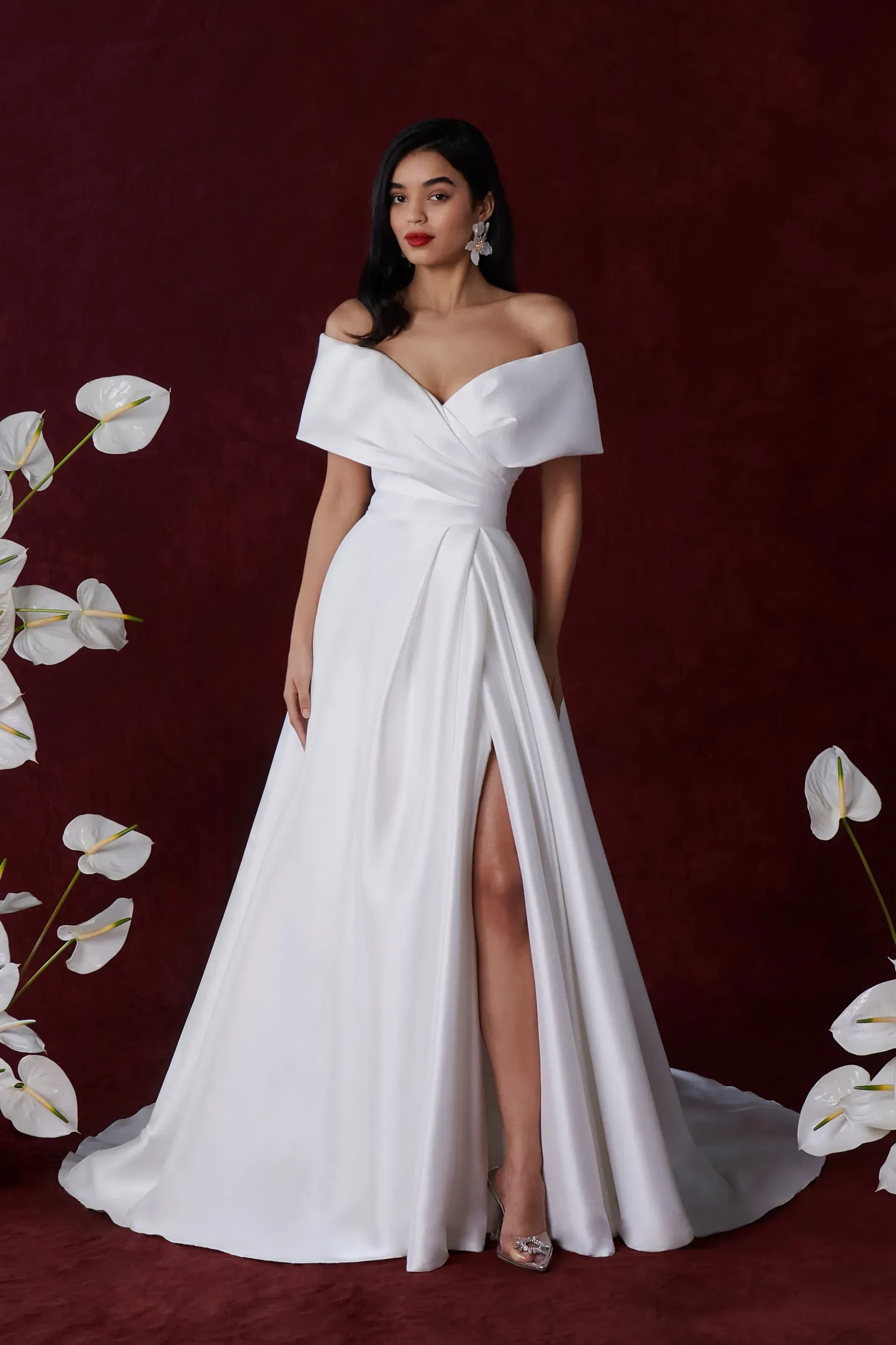 Woman in white off-the-shoulder wedding dress with a high slit, standing in front of a burgundy backdrop.