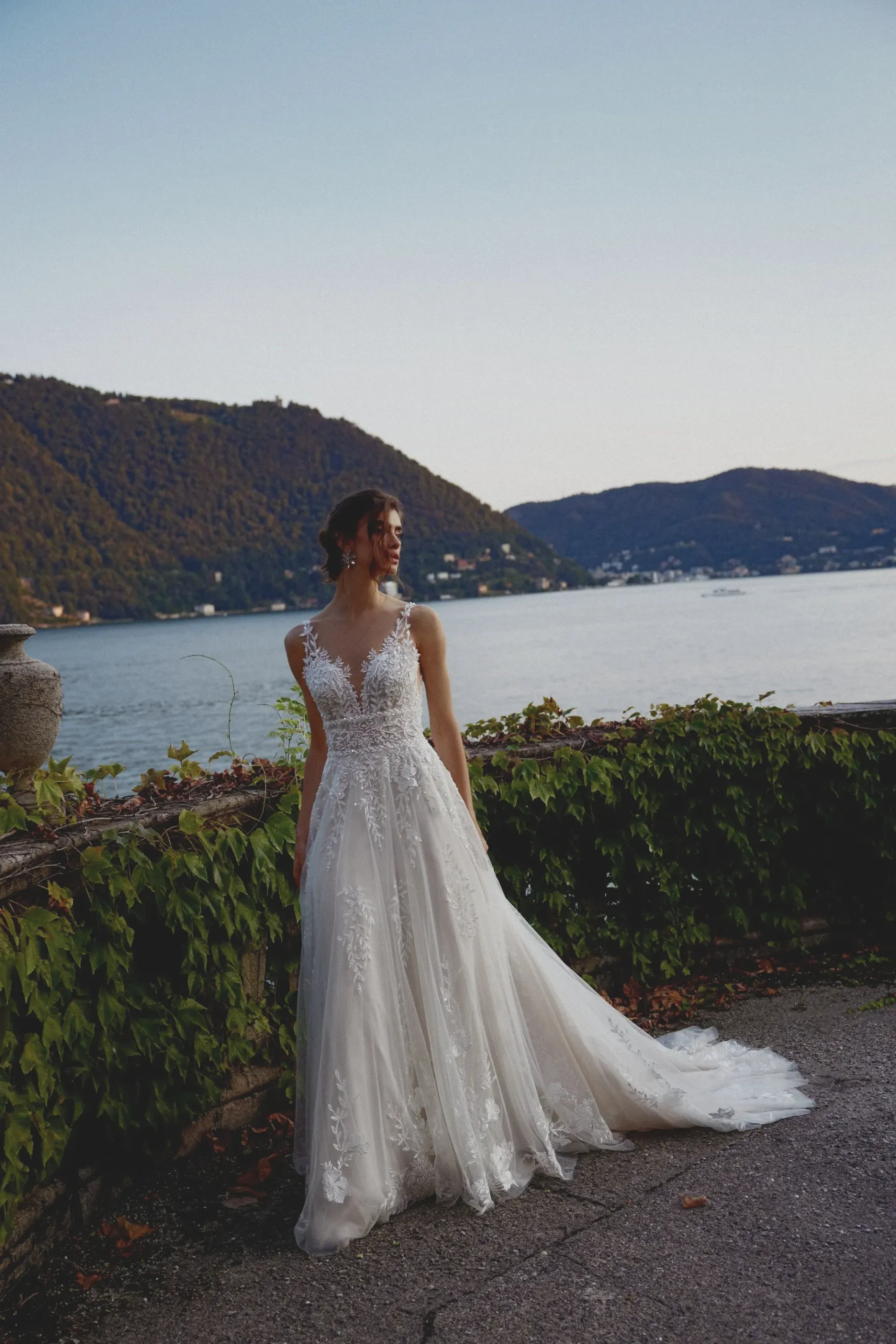 Bride in a sparkling gown, standing by a lake with mountains in the background.