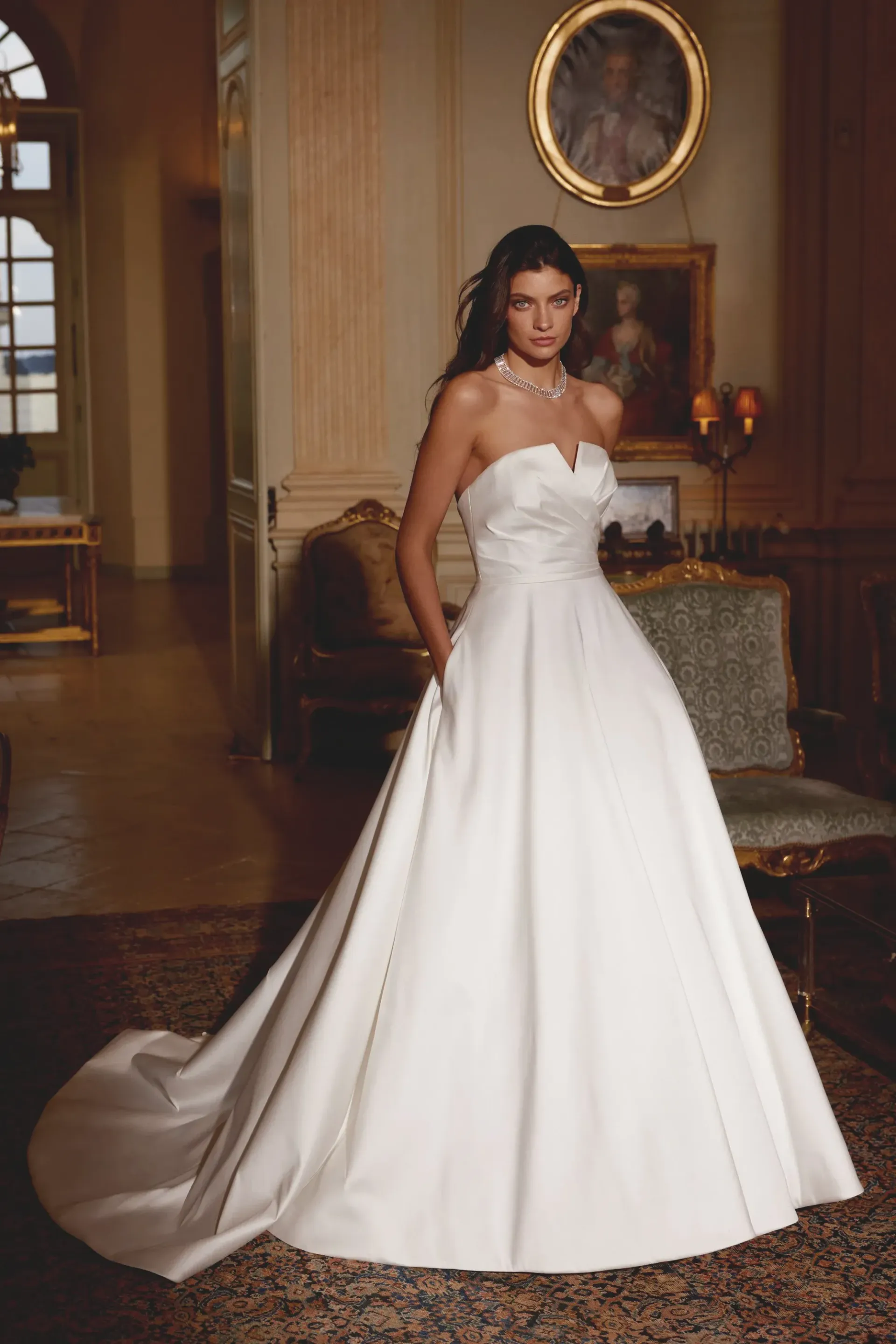 Woman in strapless white wedding dress, hands in pockets, in grand room.