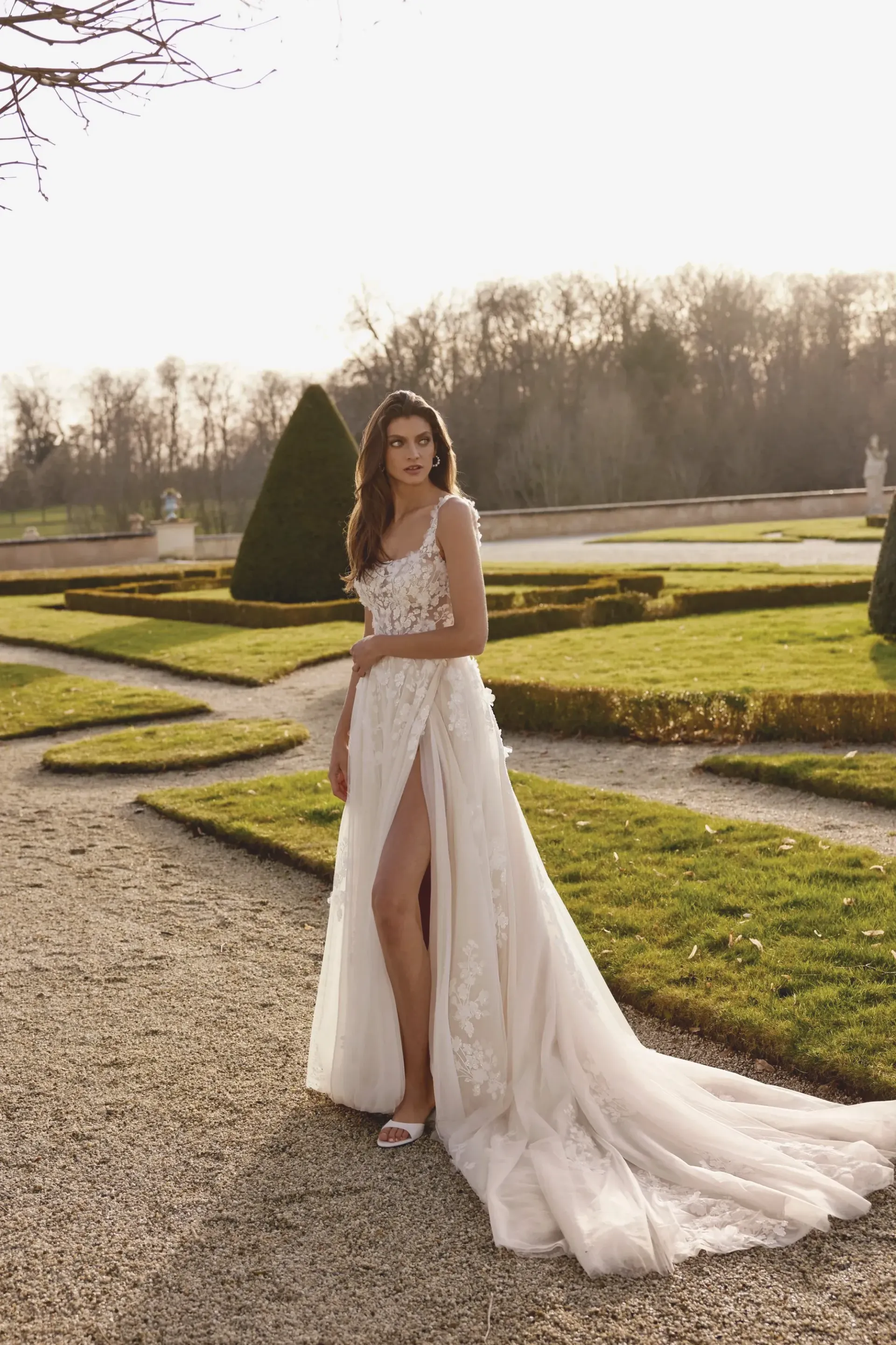 Woman in wedding dress with high slit, standing on stone path, sunny outdoor setting.