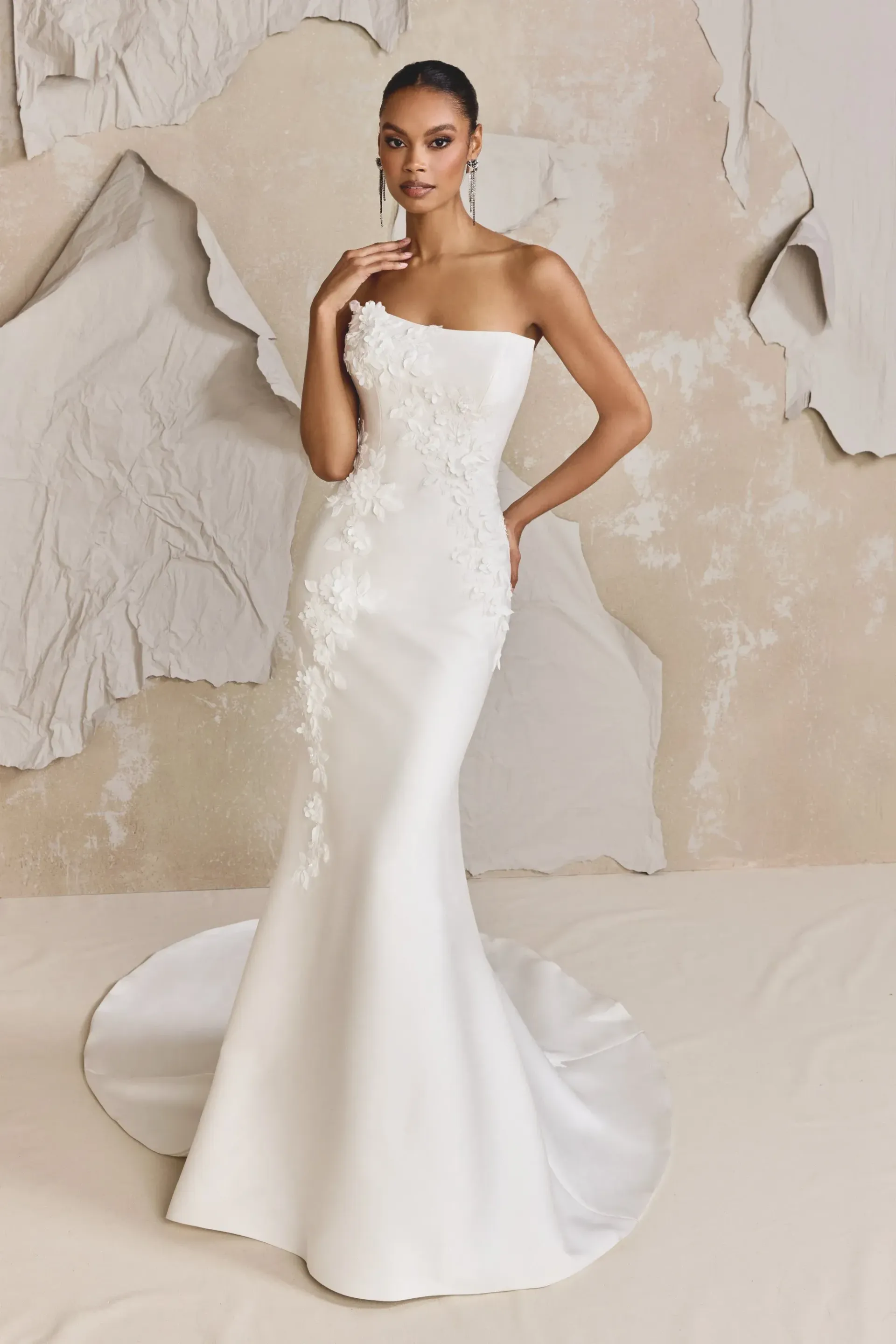Woman in a white one-shoulder wedding dress with floral embellishments, posed in a studio.