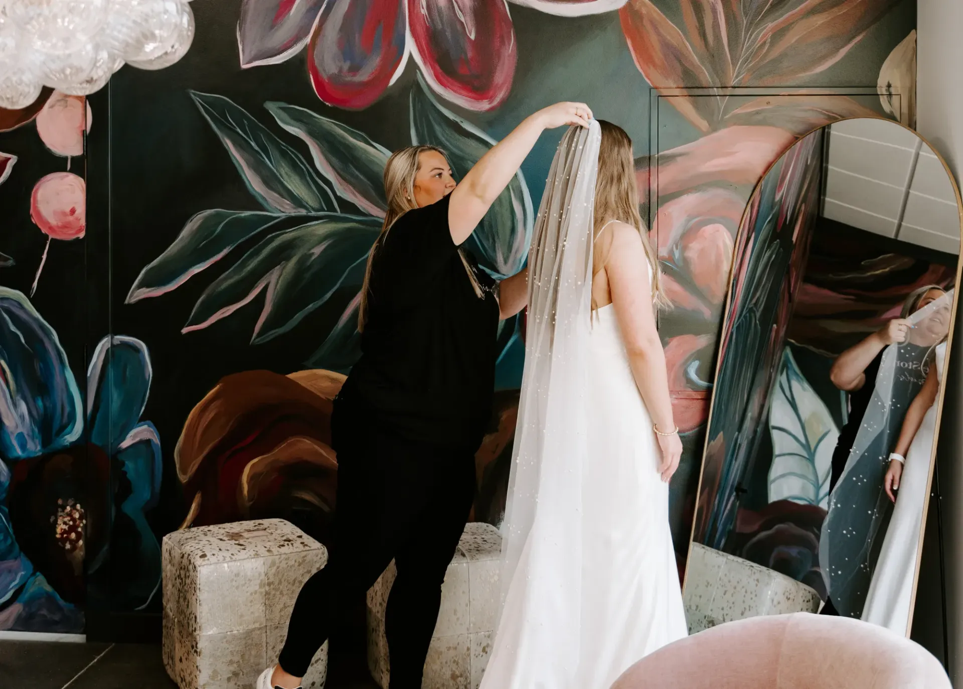 Woman in white dress, being helped with veil, by another woman in front of floral mural and mirror.