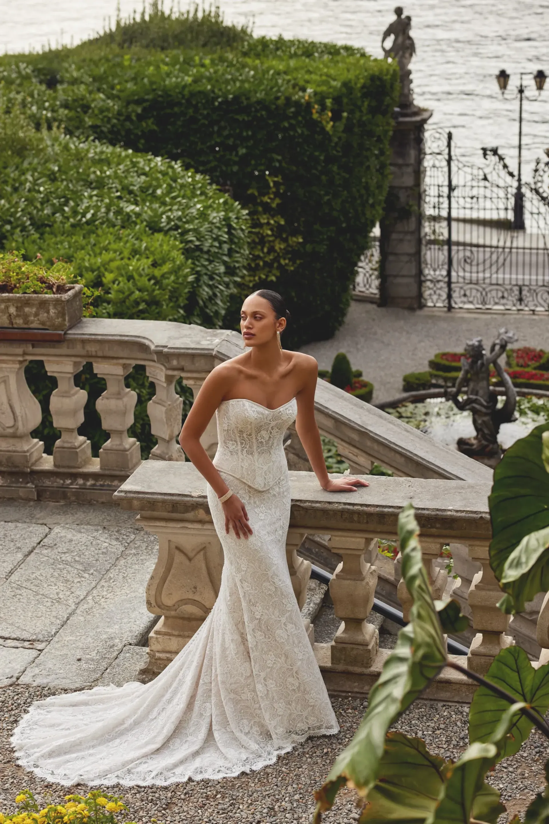Woman in a white strapless wedding dress poses on a stone balcony with a garden and water view.