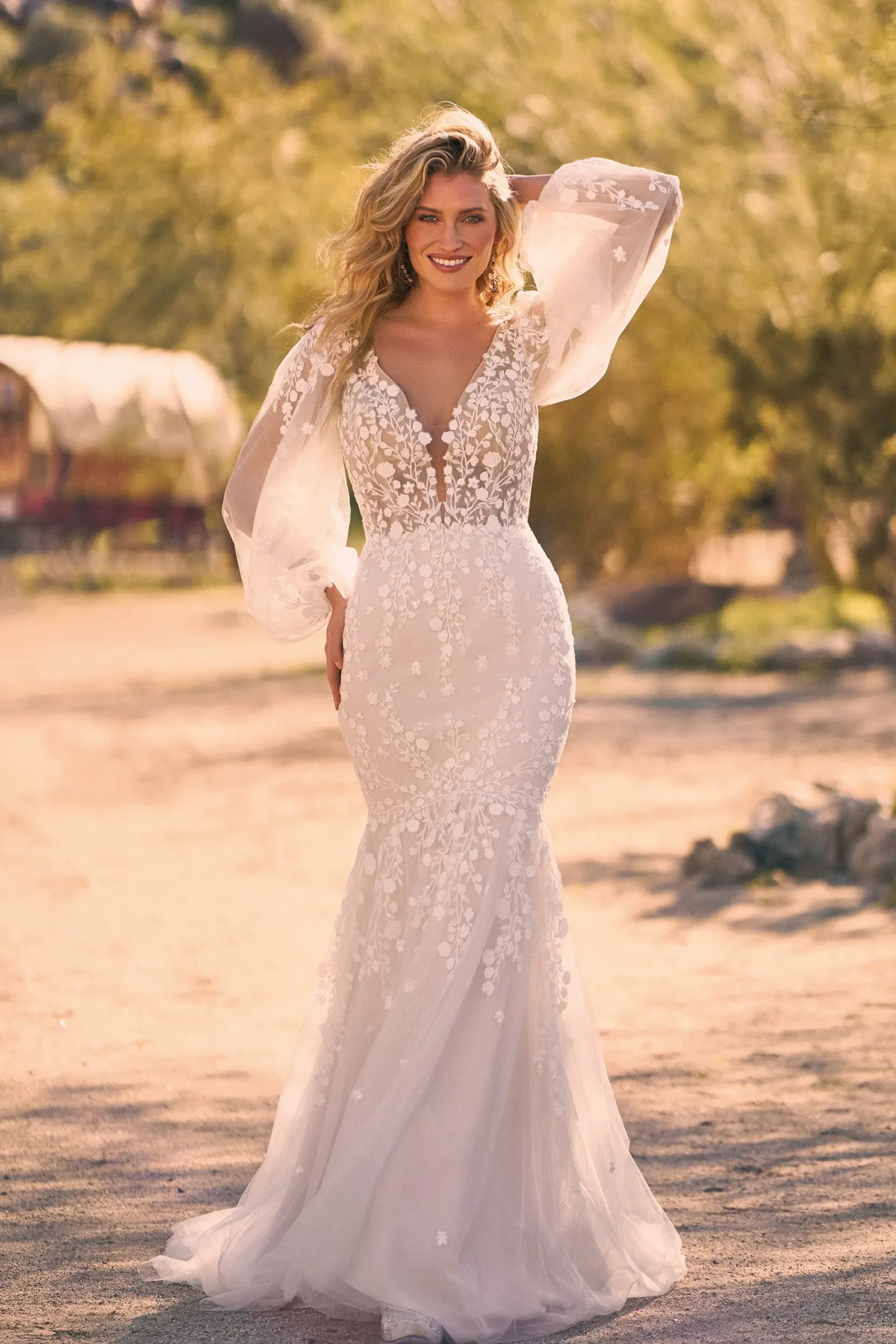 Blonde woman in a white, floral wedding dress with long sleeves, smiling outdoors.