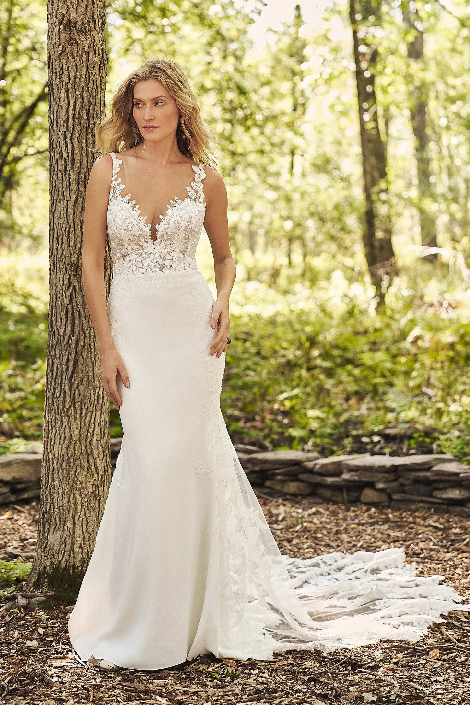 Woman in white wedding dress outdoors, leaning against tree.