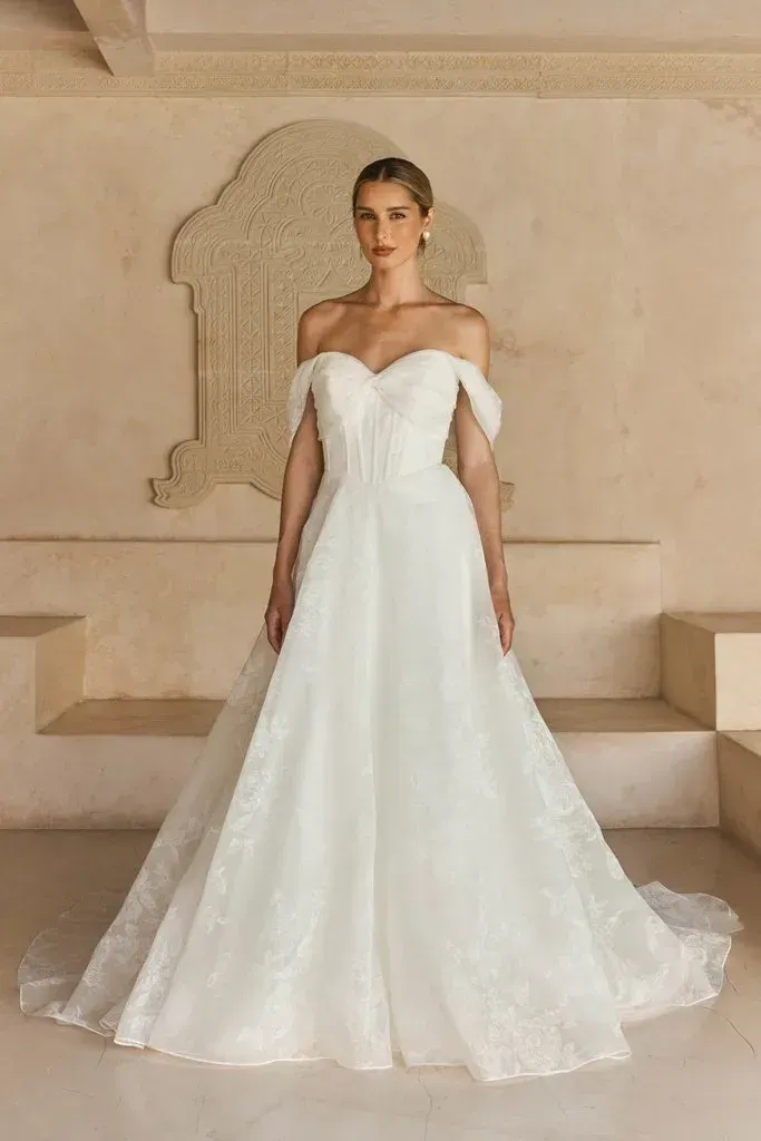 Woman in off-the-shoulder white wedding gown, standing on steps in a light-colored room.