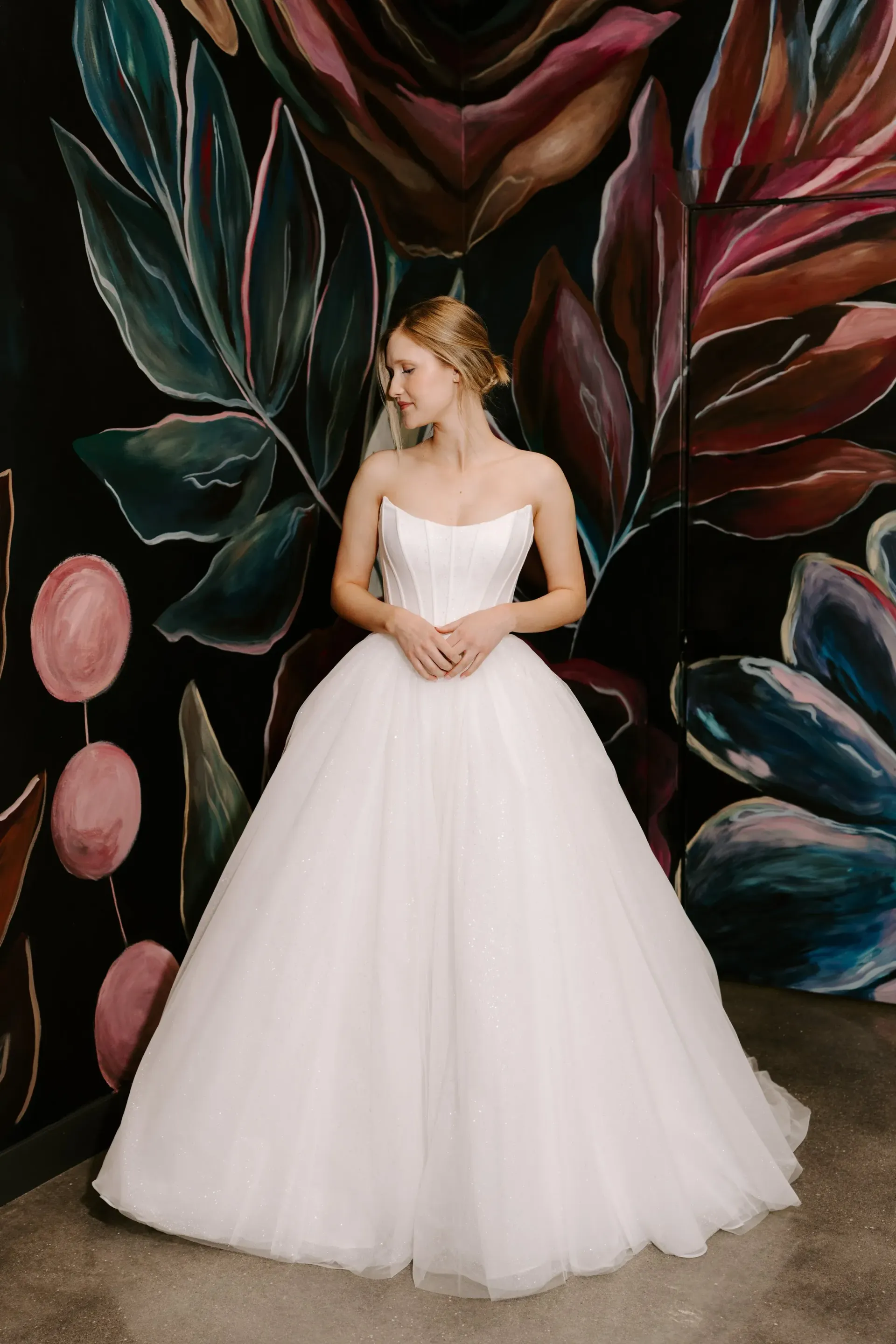 Blonde woman in a white ballgown poses against a dark wall with painted floral designs.