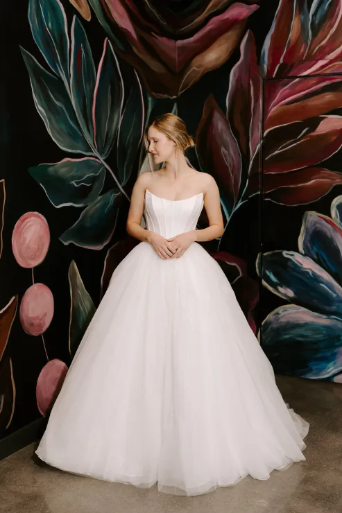 Woman in a white ballgown poses in front of a floral mural, smiling.
