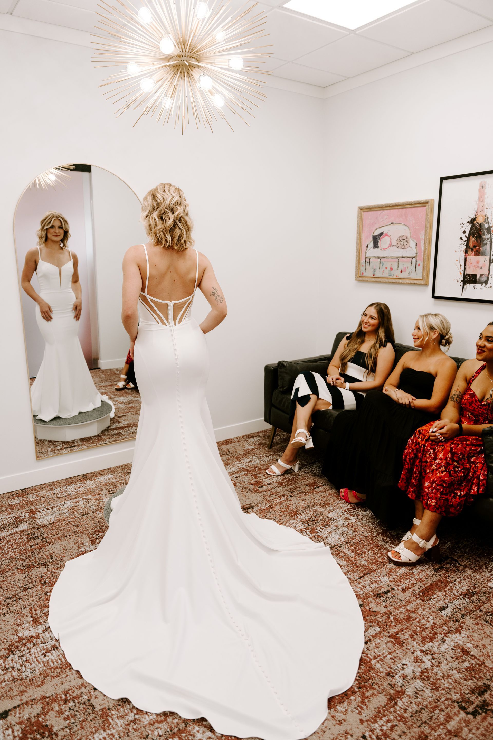 Bride in wedding dress, back to camera, in bridal shop, with friends watching.