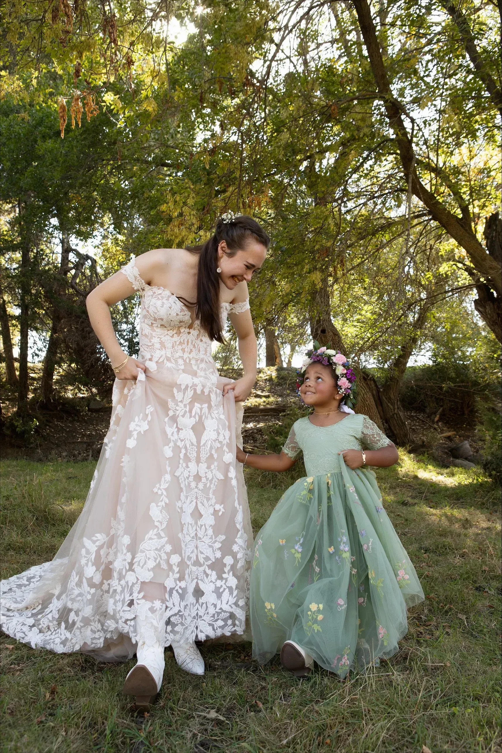Bride in lace gown and flower girl in green dress outdoors, smiling at each other.