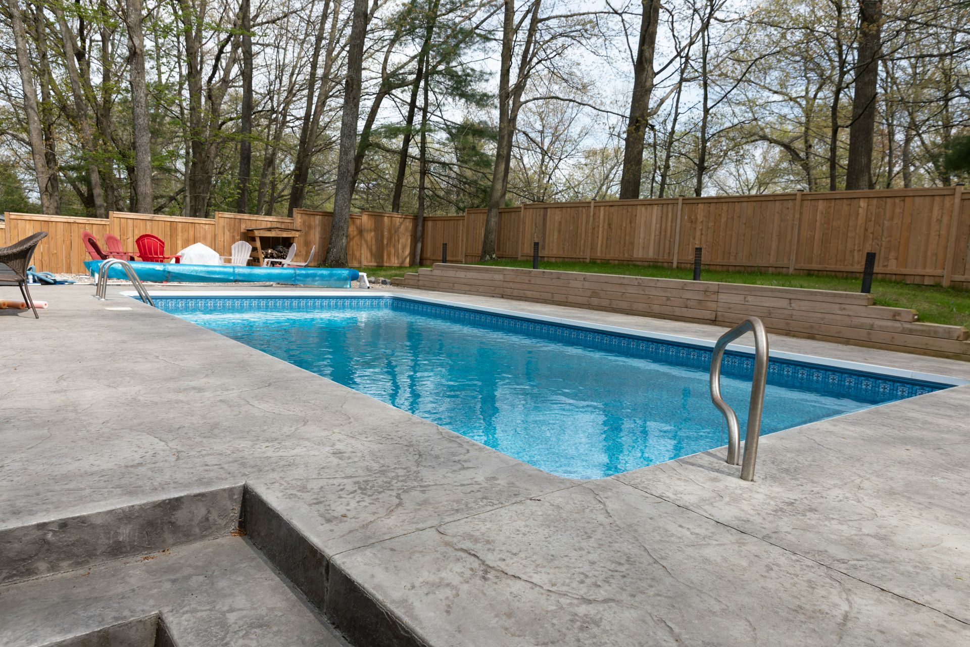 Outdoor swimming pool with blue water, concrete deck, and trees behind a wooden fence.