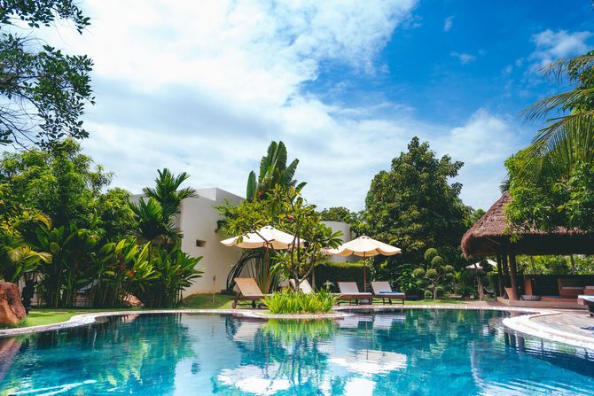 Tropical resort pool with blue water, white lounge chairs, umbrellas, and lush greenery under a bright sky