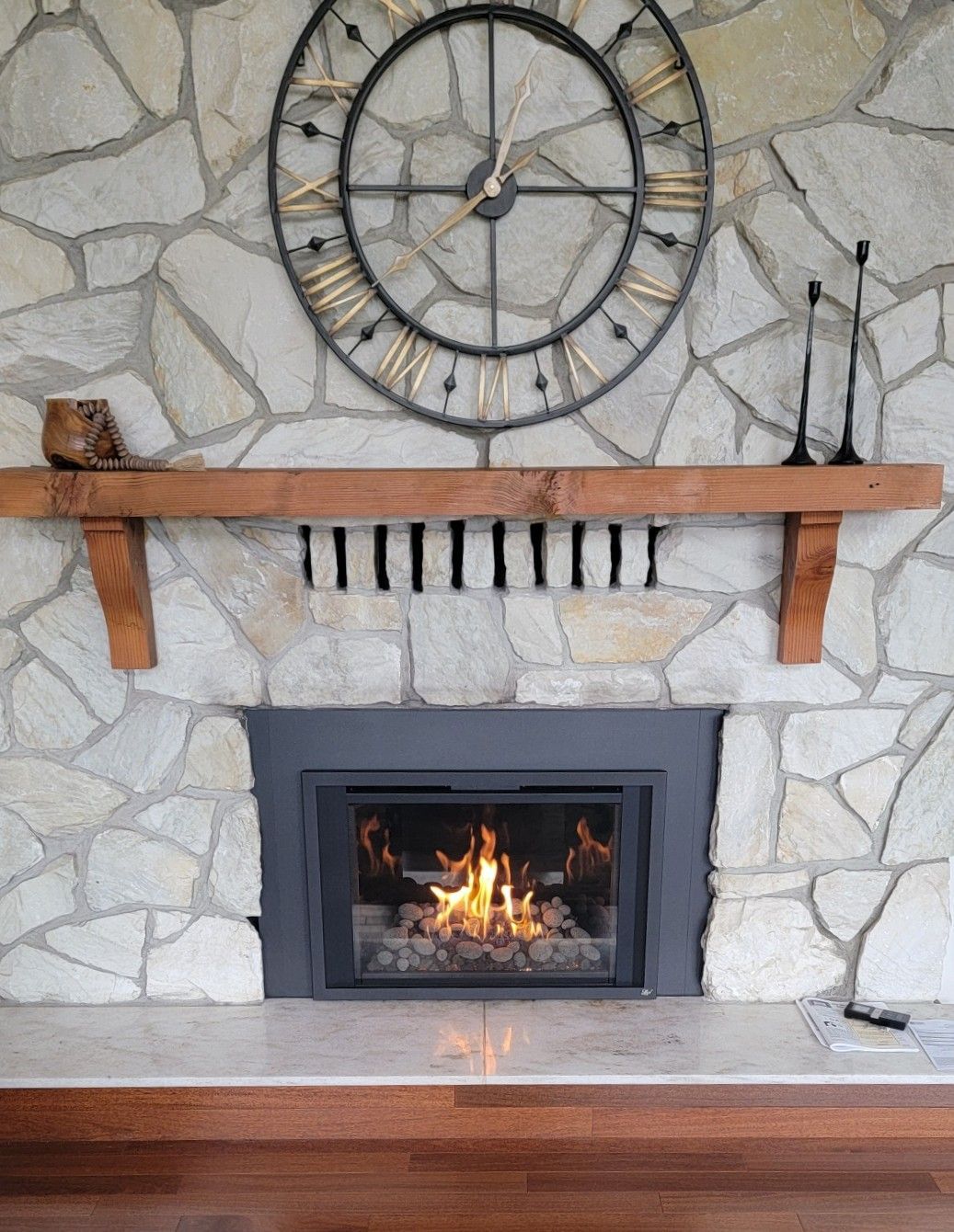 Stone fireplace with a lit fire, wooden mantel, large wall clock, and wrought-iron tools.