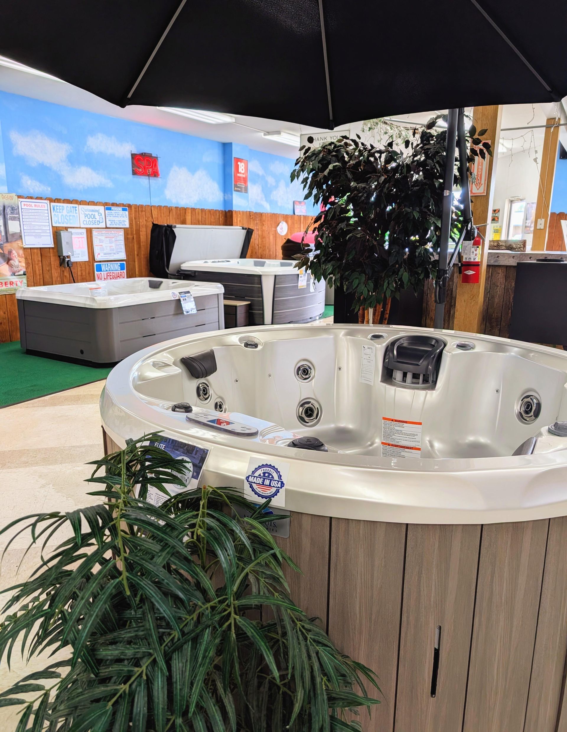 Indoor hot tub showroom displaying multiple spa models including a round white Made in USA hot tub in the foreground with jets and headrests, surrounded by decorative plants and additional spa units on display in the background. Indoor hot tub showroom displaying multiple spa models including a round white Made in USA hot tub in the foreground with jets and headrests, surrounded by decorative plants and additional spa units on display in the background.
