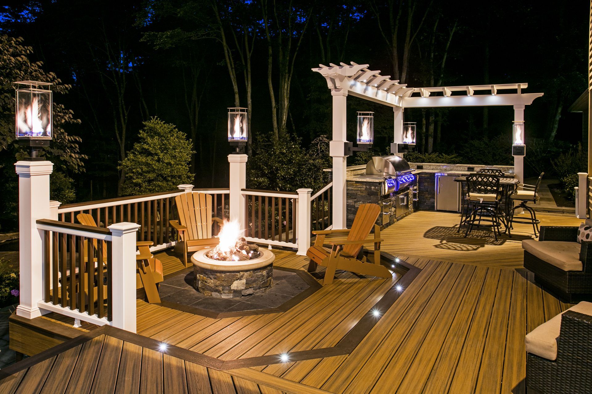 Lit wooden backyard deck with fire pit, pergola, railing lights, and outdoor seating at night