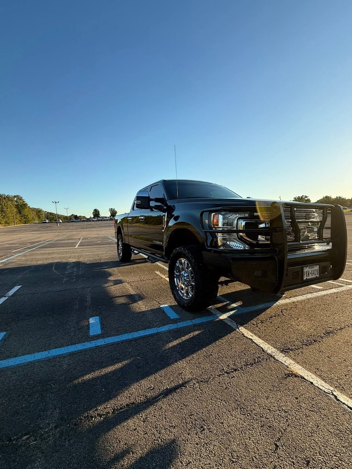 Black pickup truck in a mostly empty parking lot at sunset, casting a long shadow.