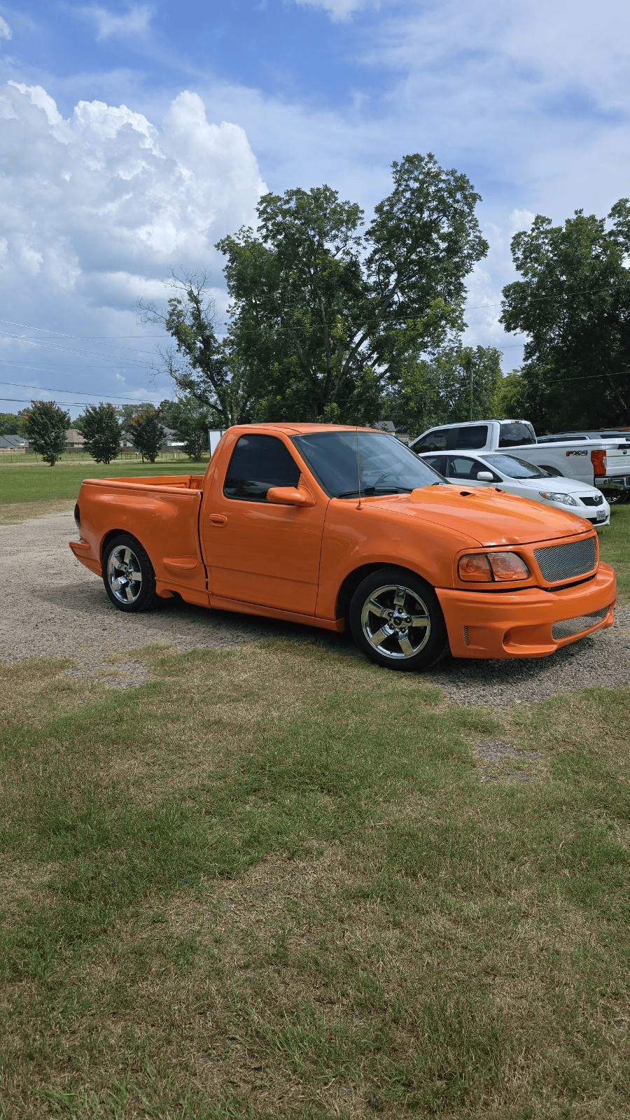 Orange pickup truck parked on grass under a cloudy sky, with trees and other cars in the background