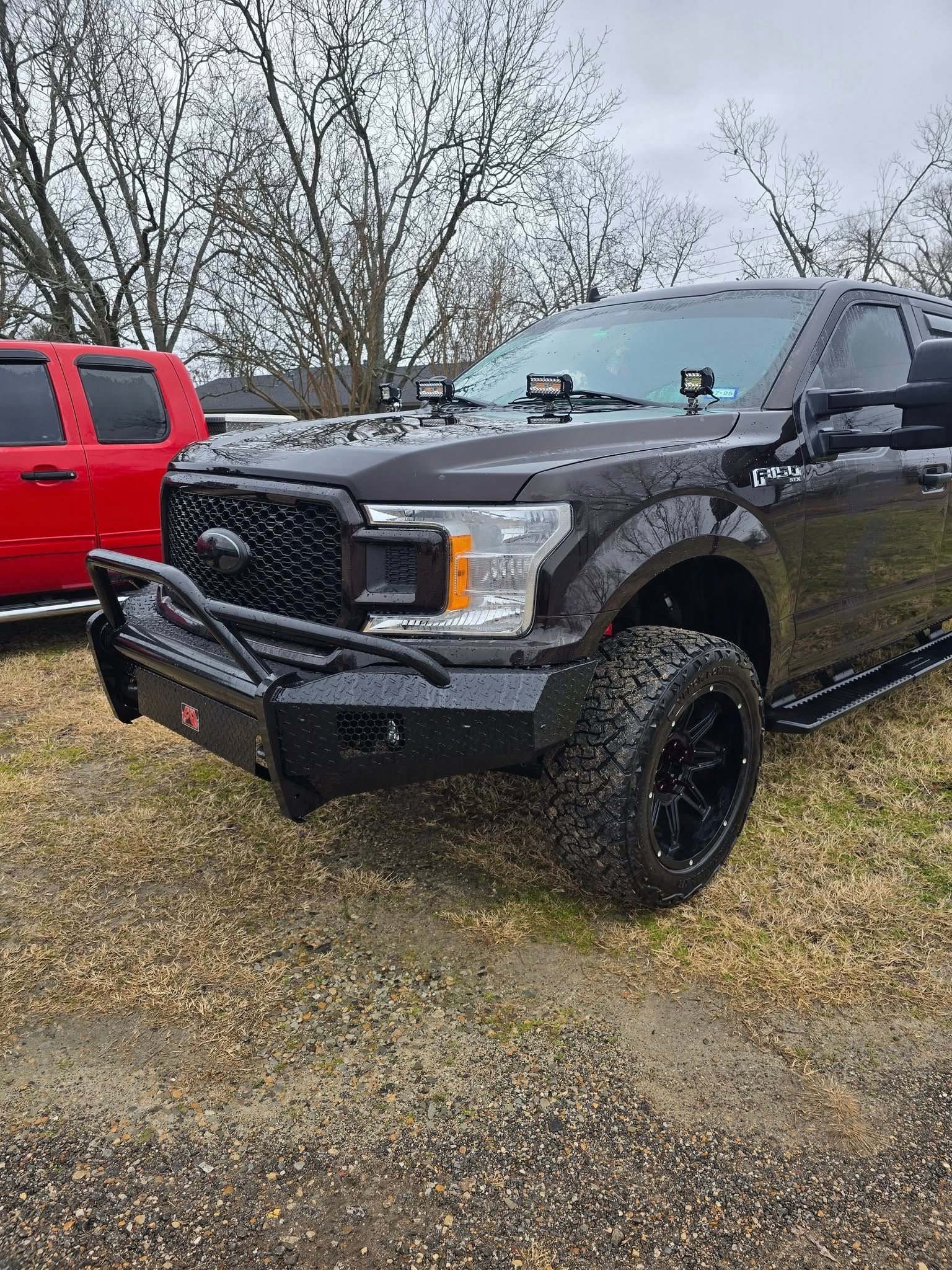 Black Ford F-150 with modified front end and black wheels, parked on grass with a red truck in the background.