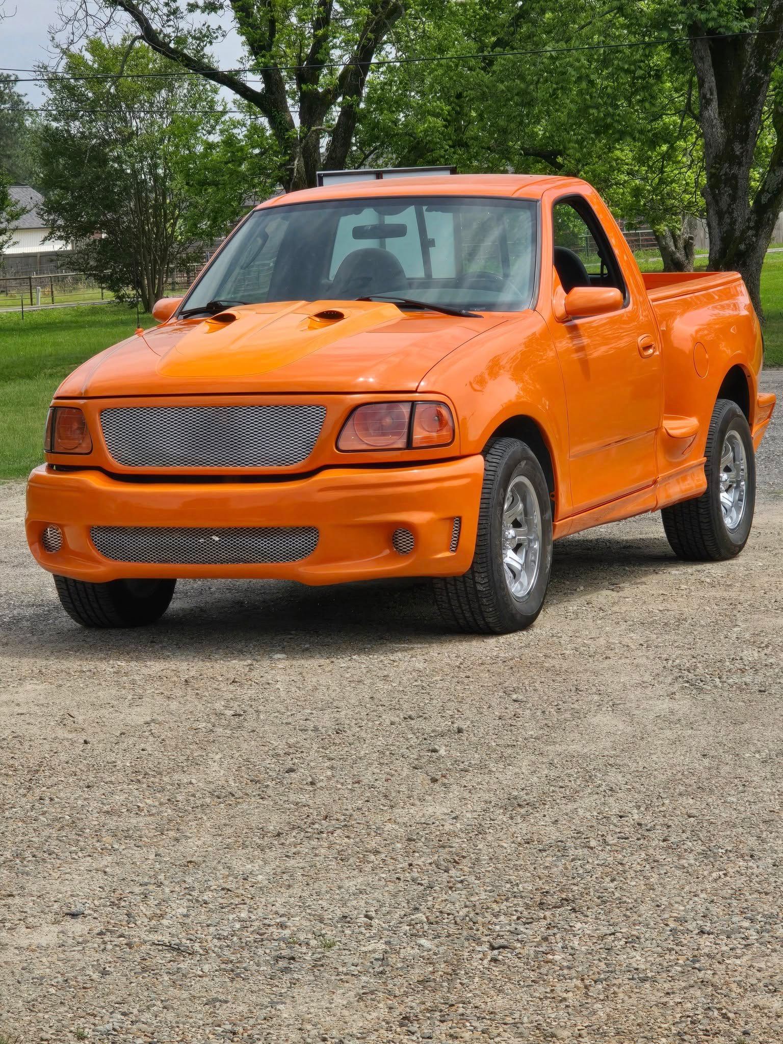 Orange Ford pickup truck with custom grill parked on gravel.