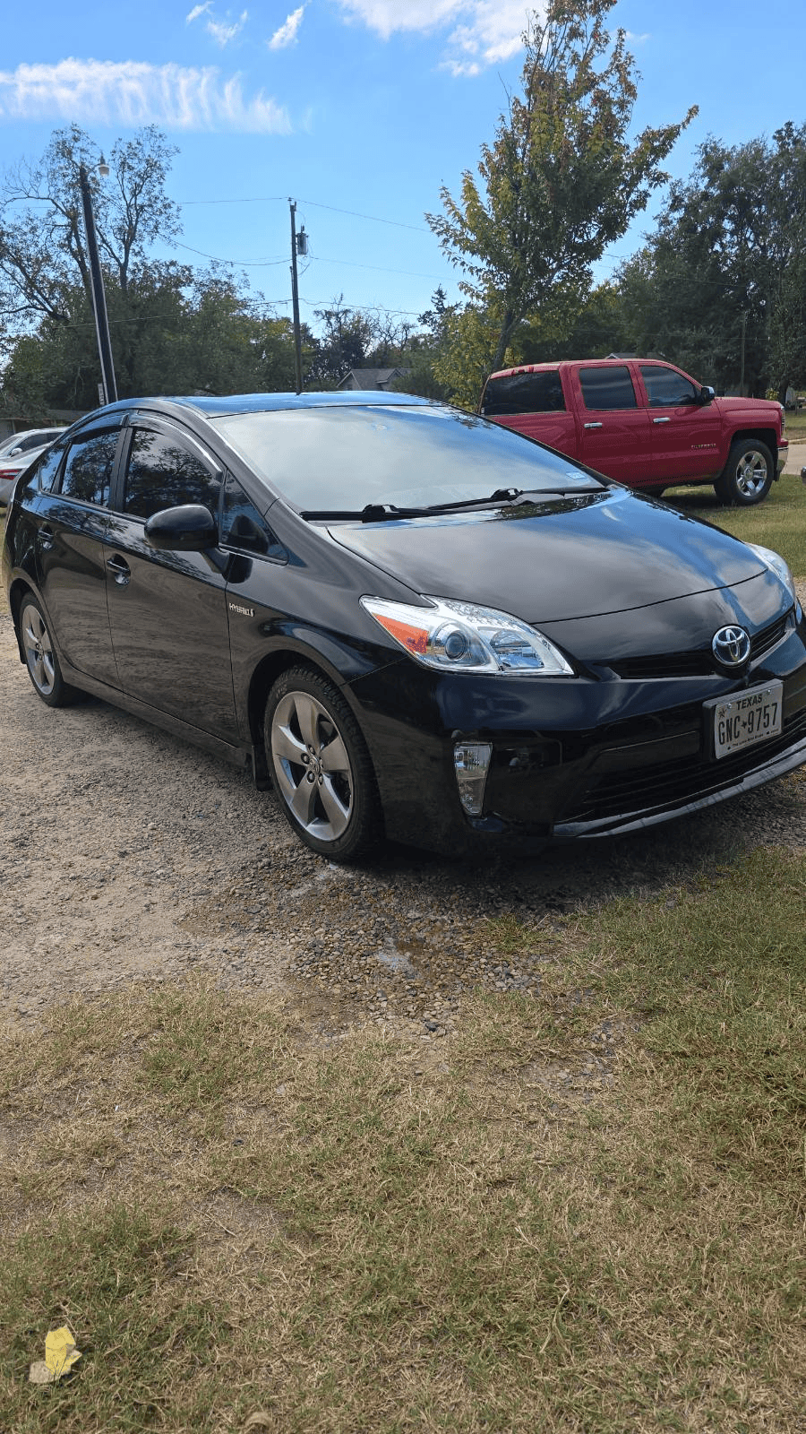 Black sedan parked on grass and gravel, with a red pickup truck behind it under a blue sky.