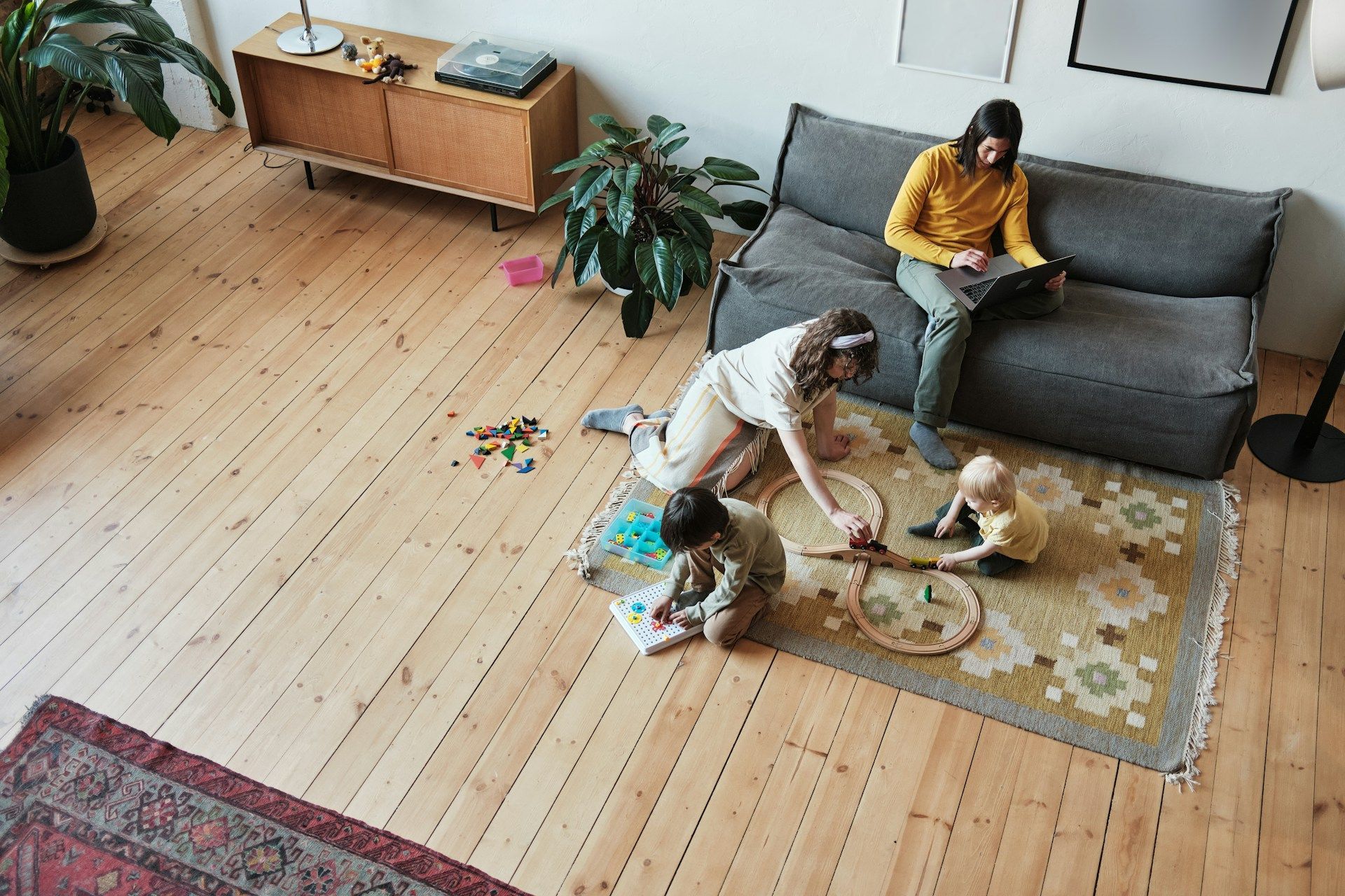 Child and adult playing on a rug in a living room with a gray sofa and wooden floor.