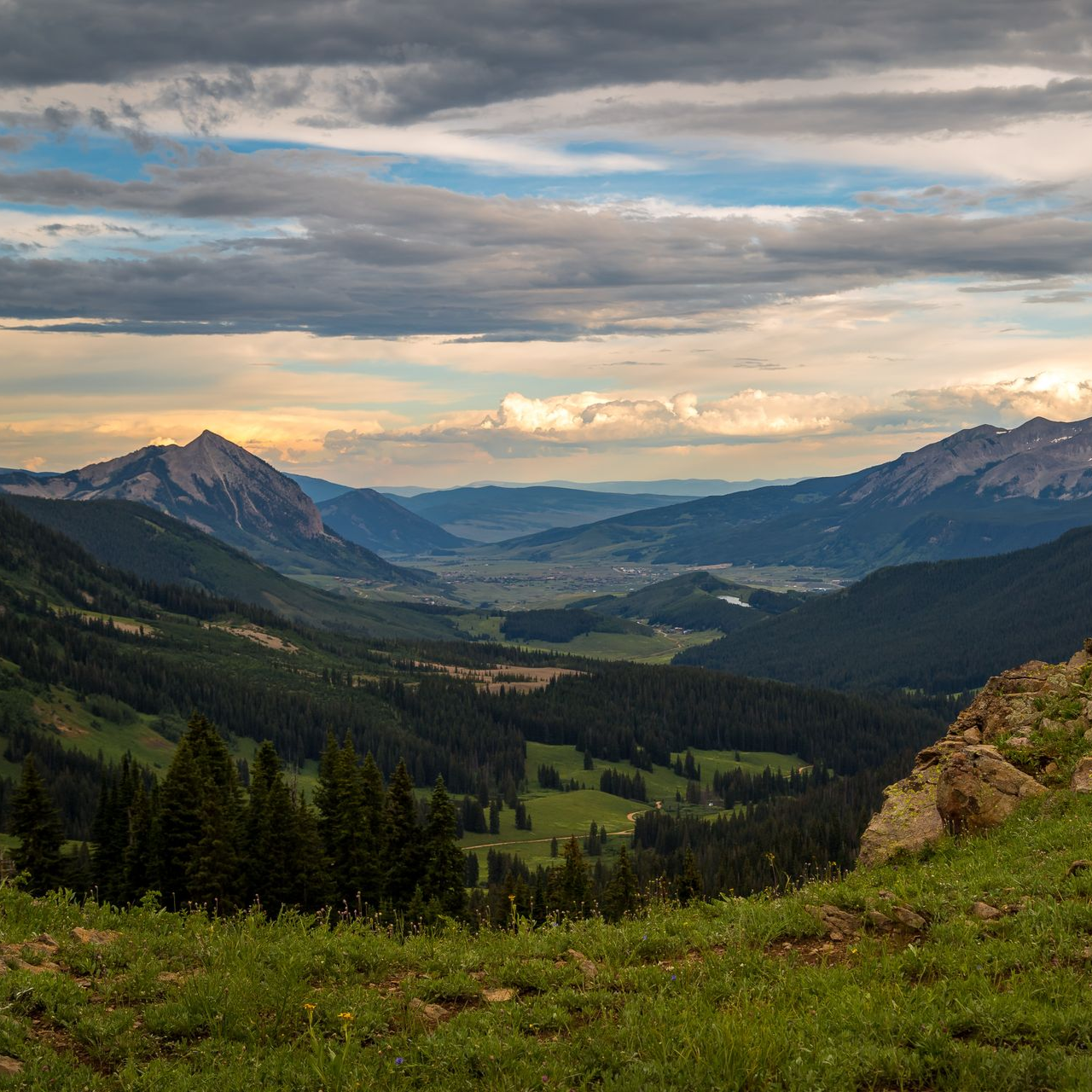Mountain valley with green slopes, forest, and cloudy sky.
