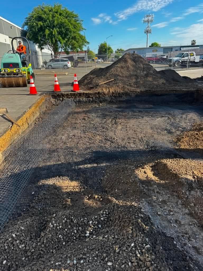 Construction site with a dirt mound, orange cones, and paved ground beside parked vehicles.
