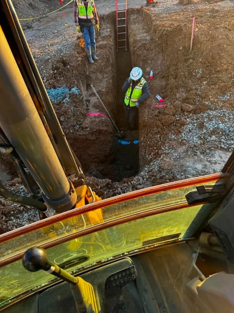 Two workers in safety vests inspect a deep trench beside heavy construction machinery.