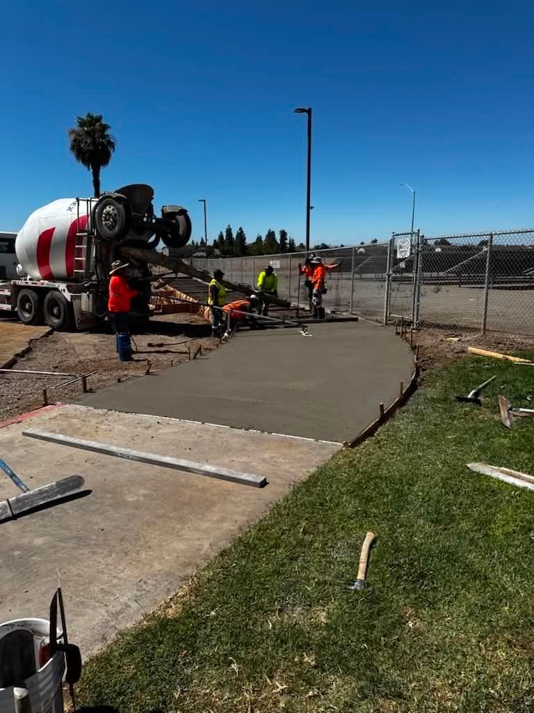 Construction workers pouring concrete walkway beside a cement truck near a fenced road site