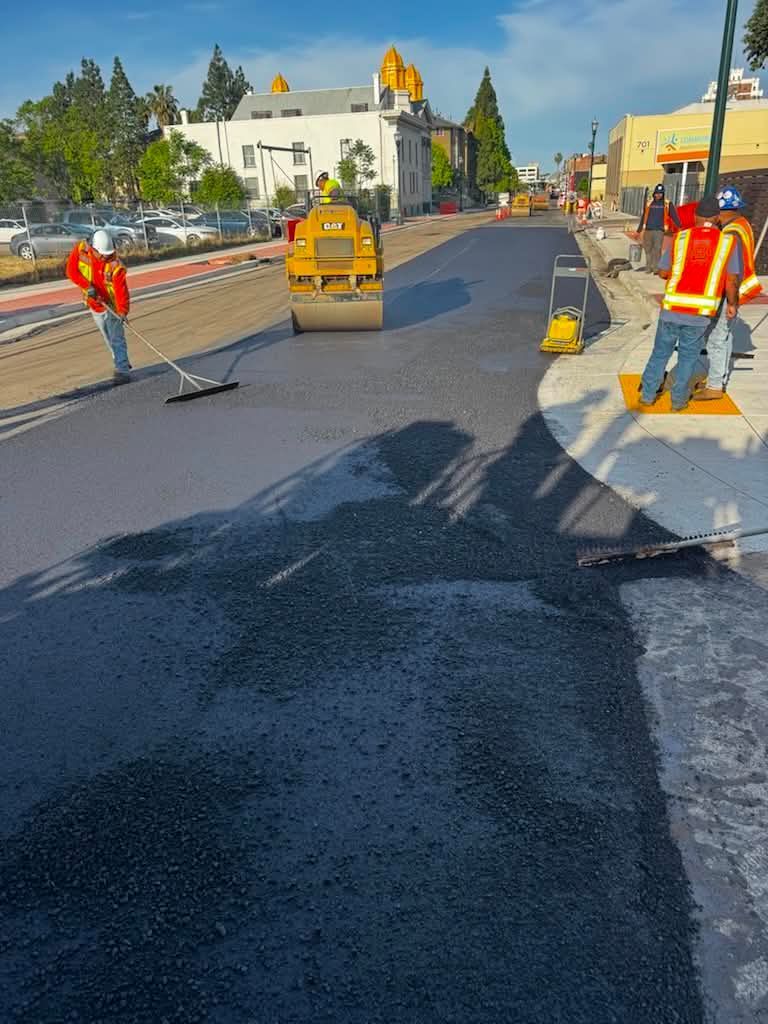 Road work crew paving a street with a roller and orange safety barriers