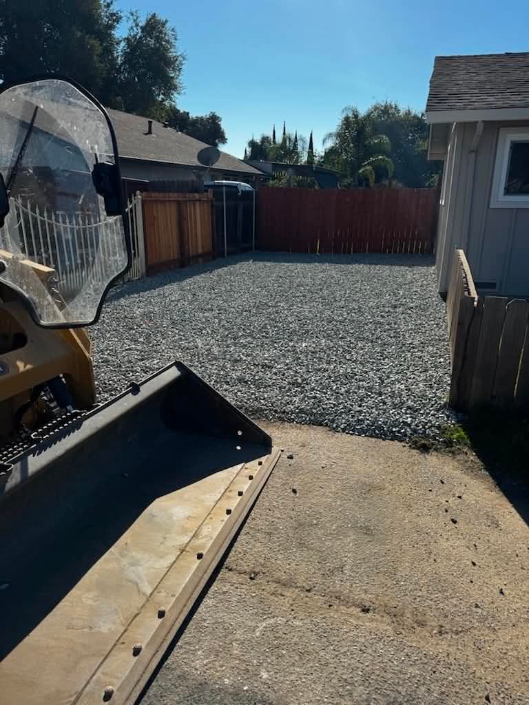 Construction equipment in a gravel yard beside a house and wooden fence.