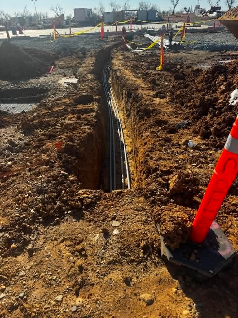 Trenches with exposed utility conduits at a construction site, marked by orange stakes and cones.